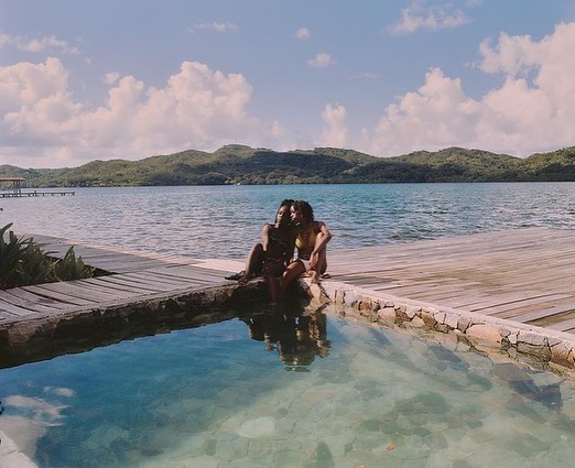 Two women sit on a stone-lined pool edge with a wooden deck and tranquil ocean view featuring distant mountains under a blue sky.