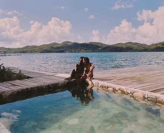 Two women sit on a stone-lined pool edge with a wooden deck and tranquil ocean view featuring distant mountains under a blue sky.
