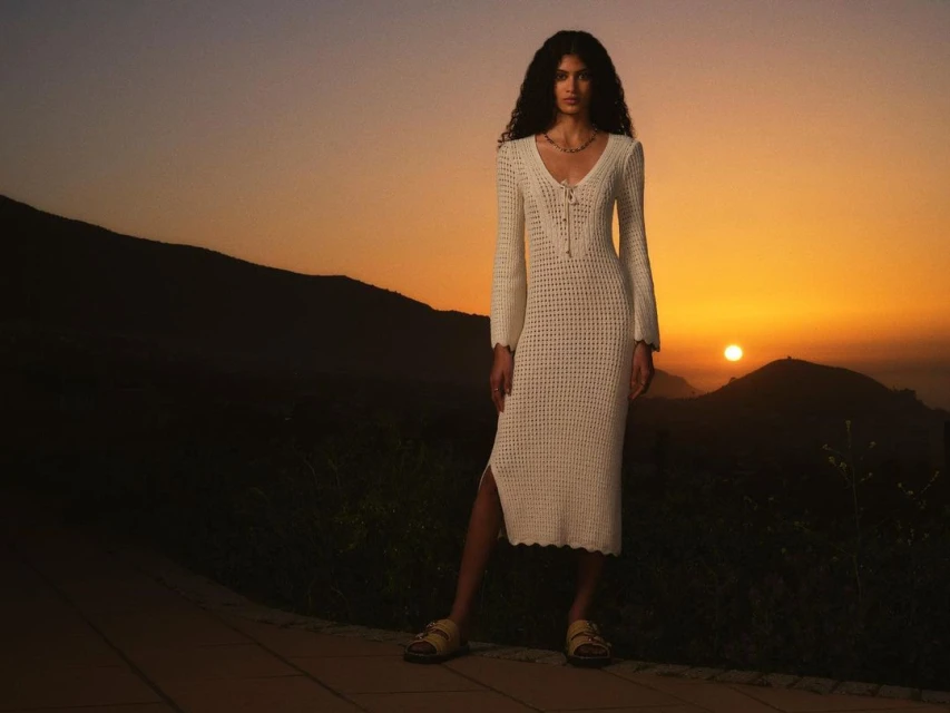 A woman in a white crochet dress poses outdoors at sunset with mountains in the background, bathed in warm, golden light.