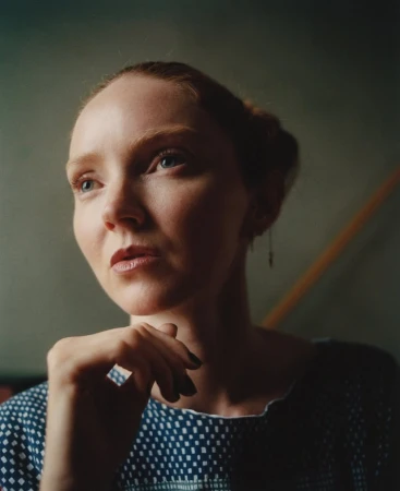 Close-up portrait of a woman with red hair resting her chin on her hand, wearing a blue patterned top against a dark wall.