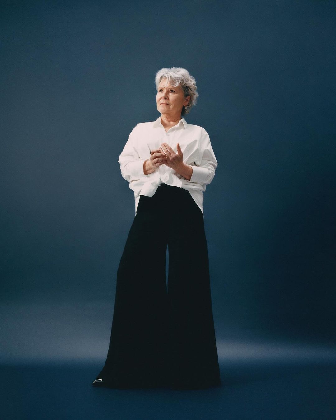 Elegant older woman in white shirt and black palazzo pants poses in a full shot against a dark teal backdrop, looking off-camera.
