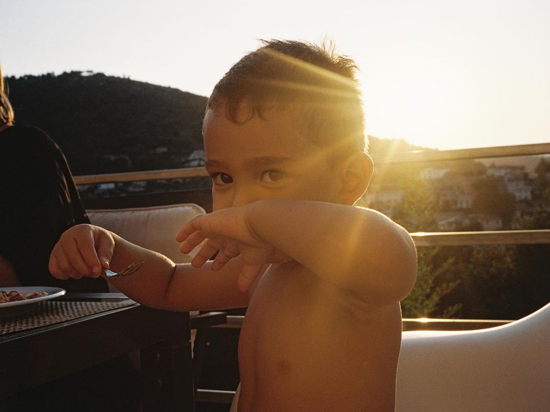 Young child holding up a fork while seated outdoors at sunset with hills visible in the background.