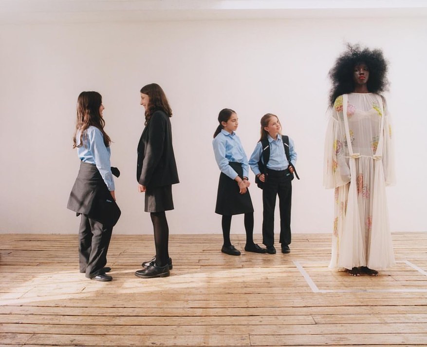 Fashion shot featuring an adult female wearing a sheer, floral print dress standing in front of three schoolgirls in uniform.