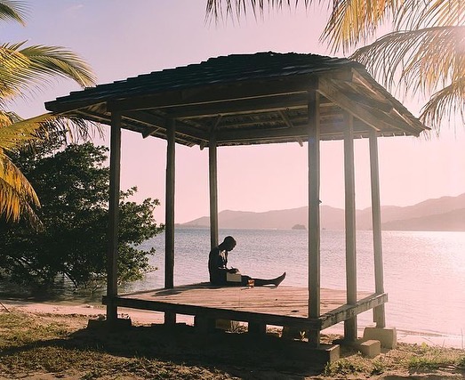 Person sitting in a gazebo overlooking ocean with tropical foliage, mountains, and a light pink sky, creating a serene and tranquil scene.