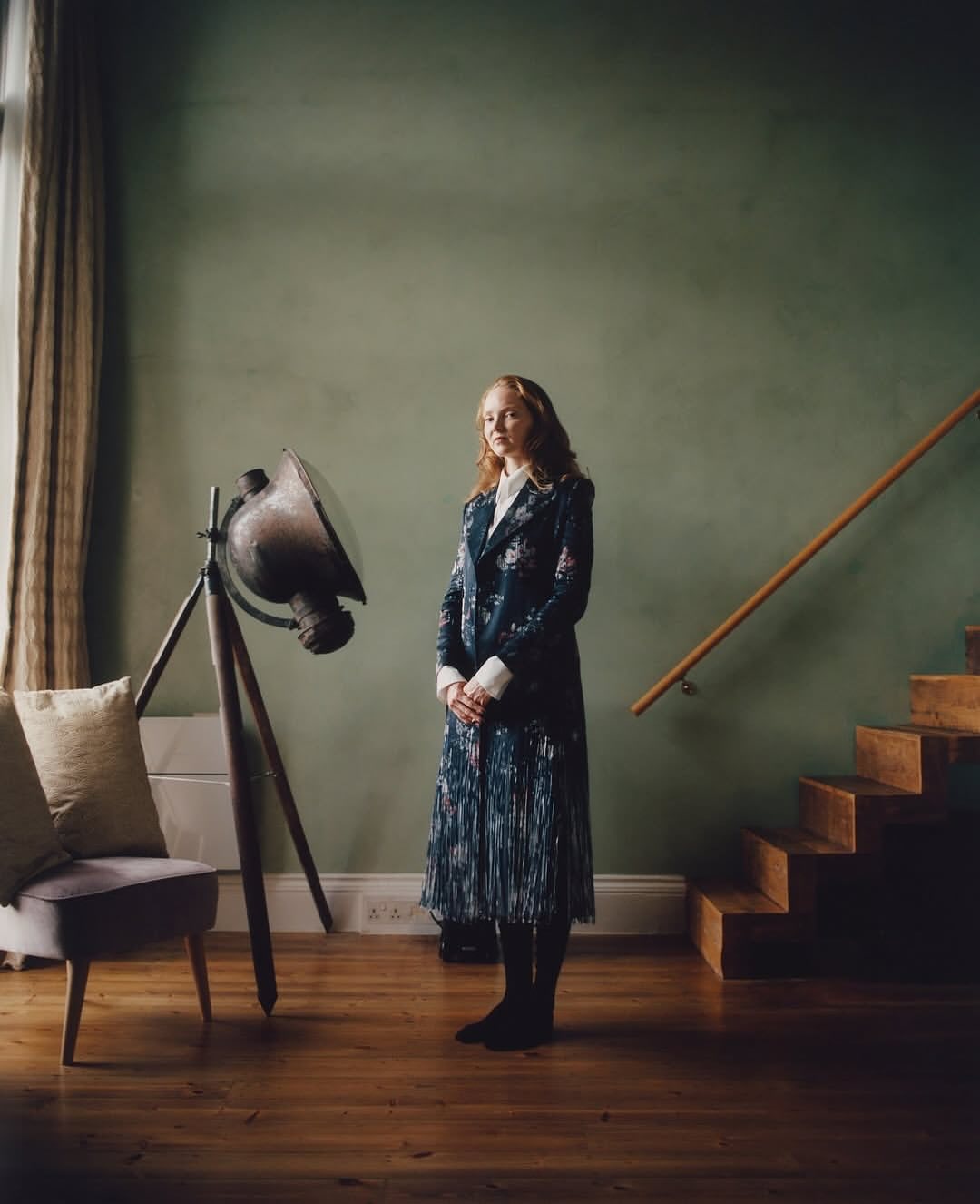 Portrait of an adult woman in a floral coat standing indoors near a vintage lamp, wood stairs, and a soft green wall.