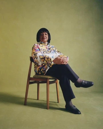 An older Black woman seated on a wooden chair wearing a patterned blouse and dark trousers against an olive green backdrop.
