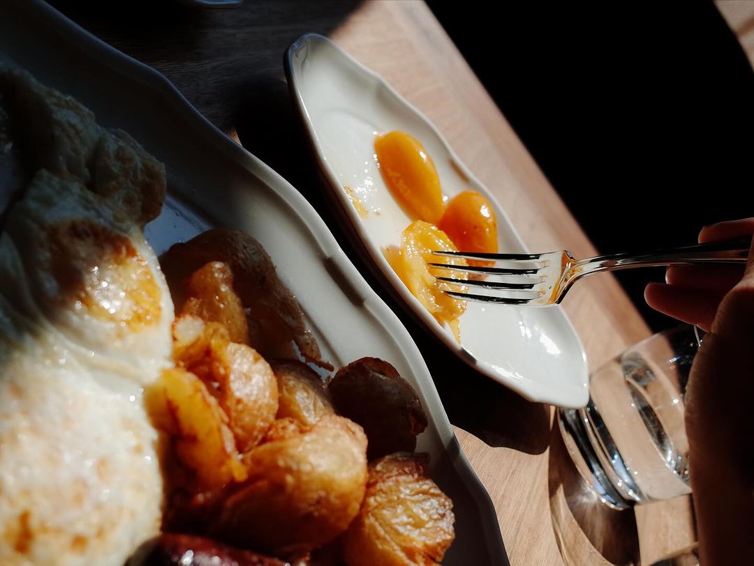 Overhead shot of a breakfast with fried eggs, potatoes, sausage, and fruit being eaten with a fork on a wooden table.