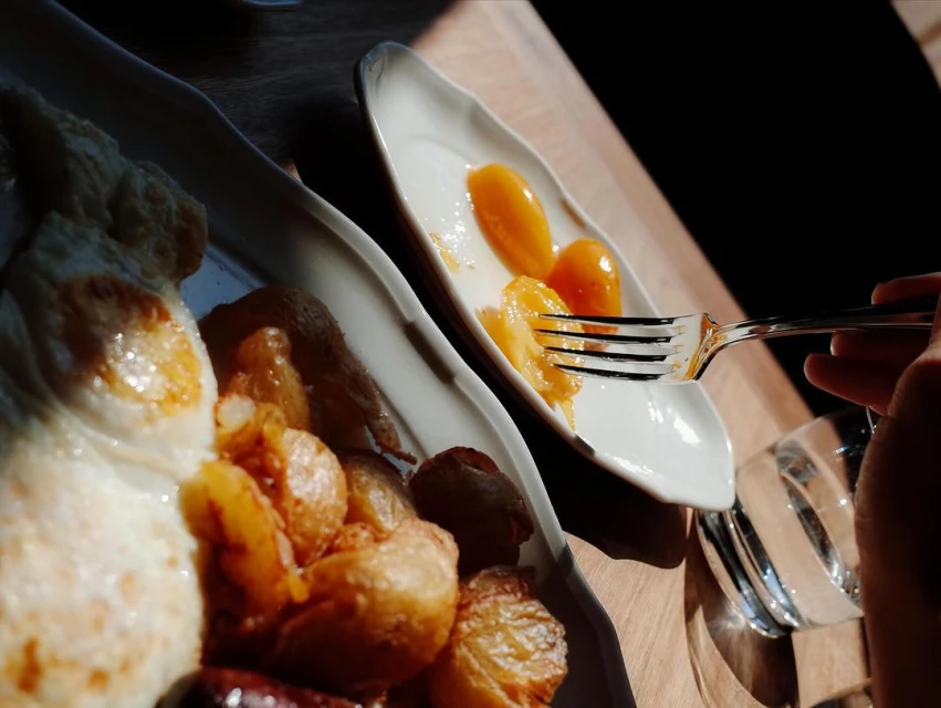 Overhead shot of a breakfast with fried eggs, potatoes, sausage, and fruit being eaten with a fork on a wooden table.