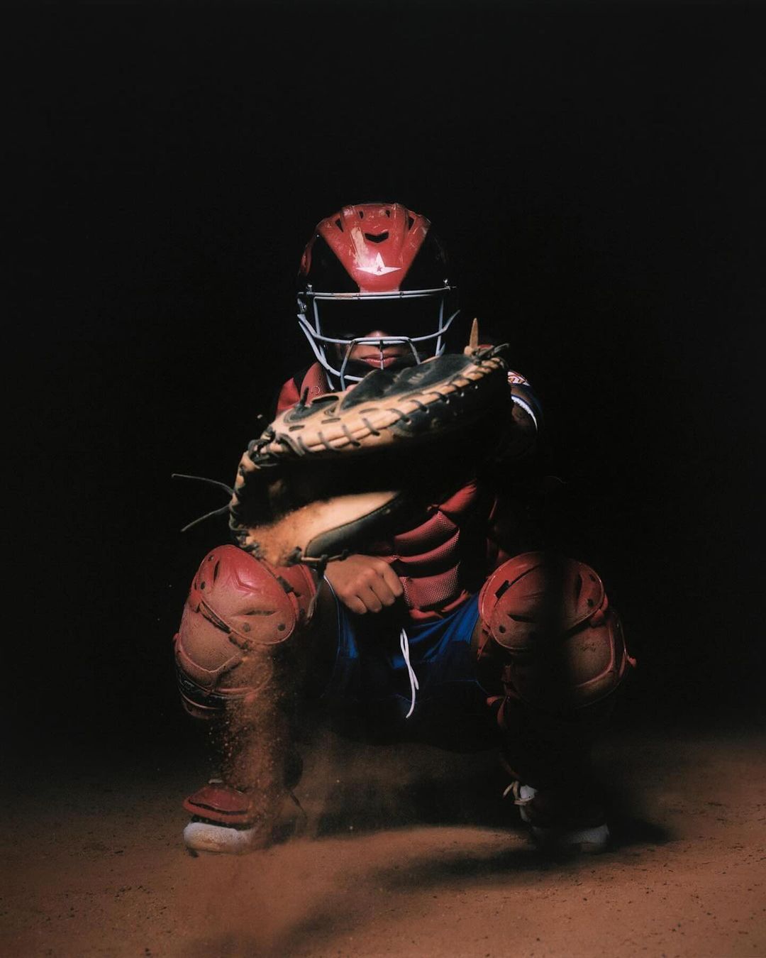 A baseball catcher in full protective gear, squatting on a dirt field, ready to catch the ball in a dimly lit indoor sporting environment.