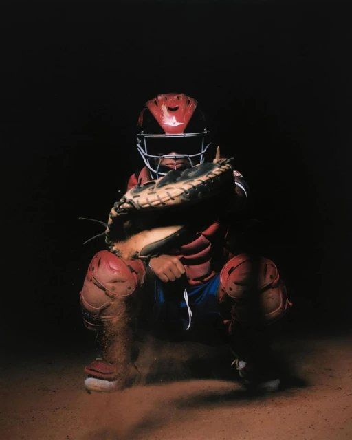A baseball catcher in full protective gear, squatting on a dirt field, ready to catch the ball in a dimly lit indoor sporting environment.