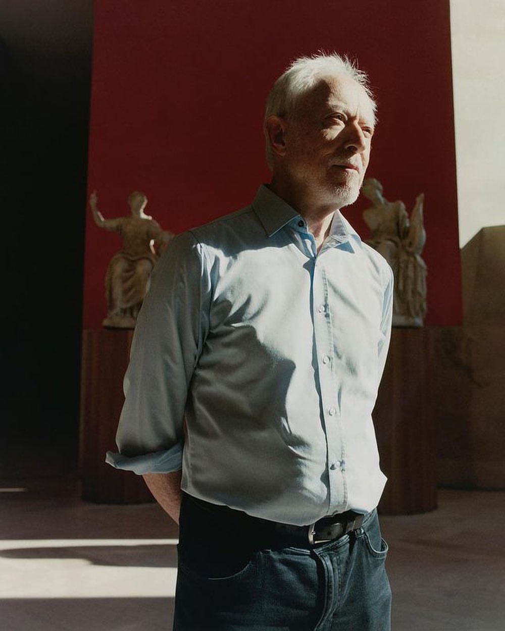 Mature man stands in a light-filled interior against a red wall with statues. He wears a light-blue shirt, jeans, and a black belt.