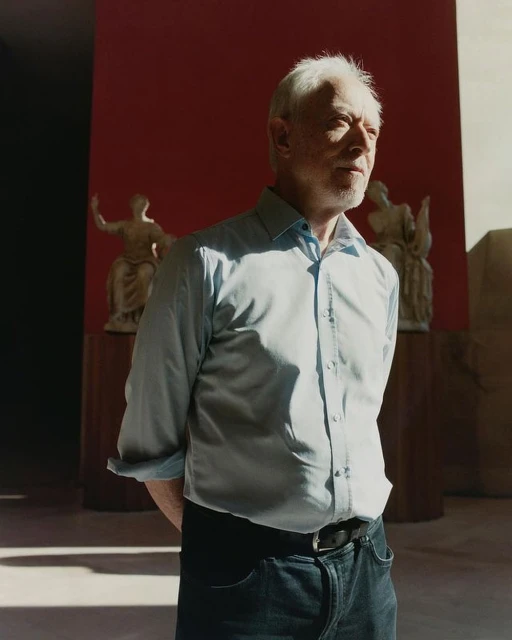 Mature man stands in a light-filled interior against a red wall with statues. He wears a light-blue shirt, jeans, and a black belt.