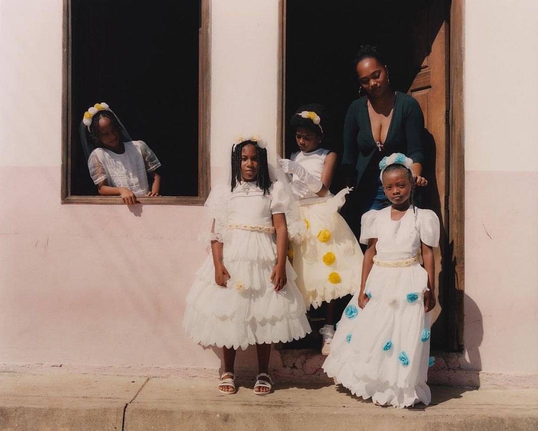 Group of young Black girls in formal dresses pose with an adult female outside a light pink building with windows and a doorway.