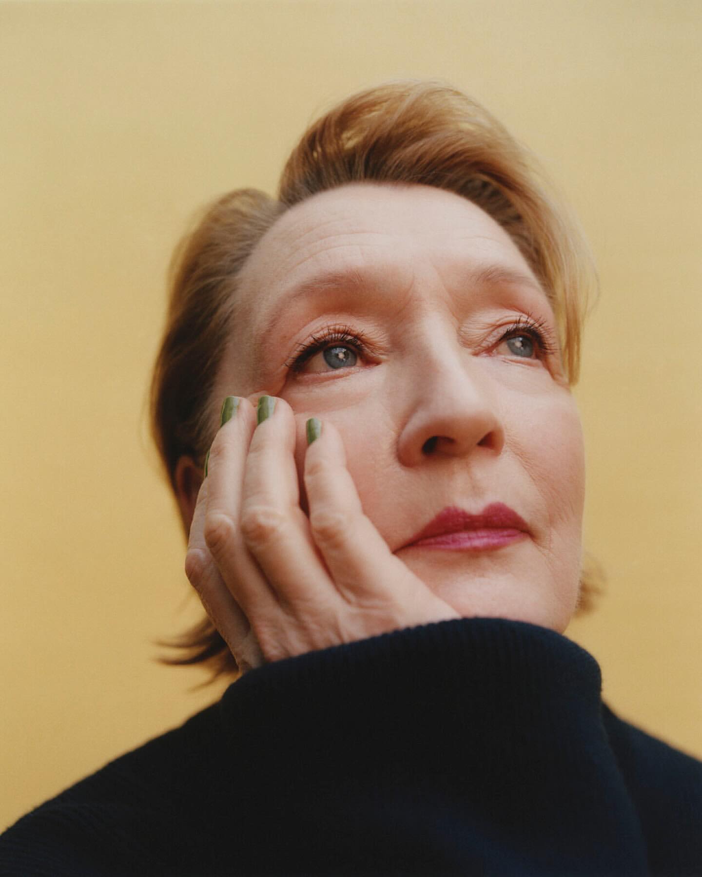 Close-up portrait of an adult woman with short red hair and green nails, resting her face on her hand against a solid yellow background.