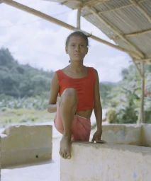 Young child in a red top and pink shorts standing outdoors with lush green hills and a corrugated roof shelter visible in the background.