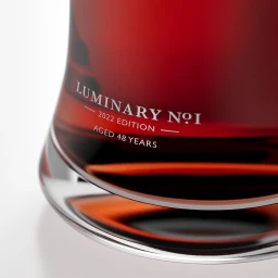 Close-up of a dark amber glass bottle base with white text reading Luminary No 1 2022 Edition Aged 48 Years against a white backdrop.