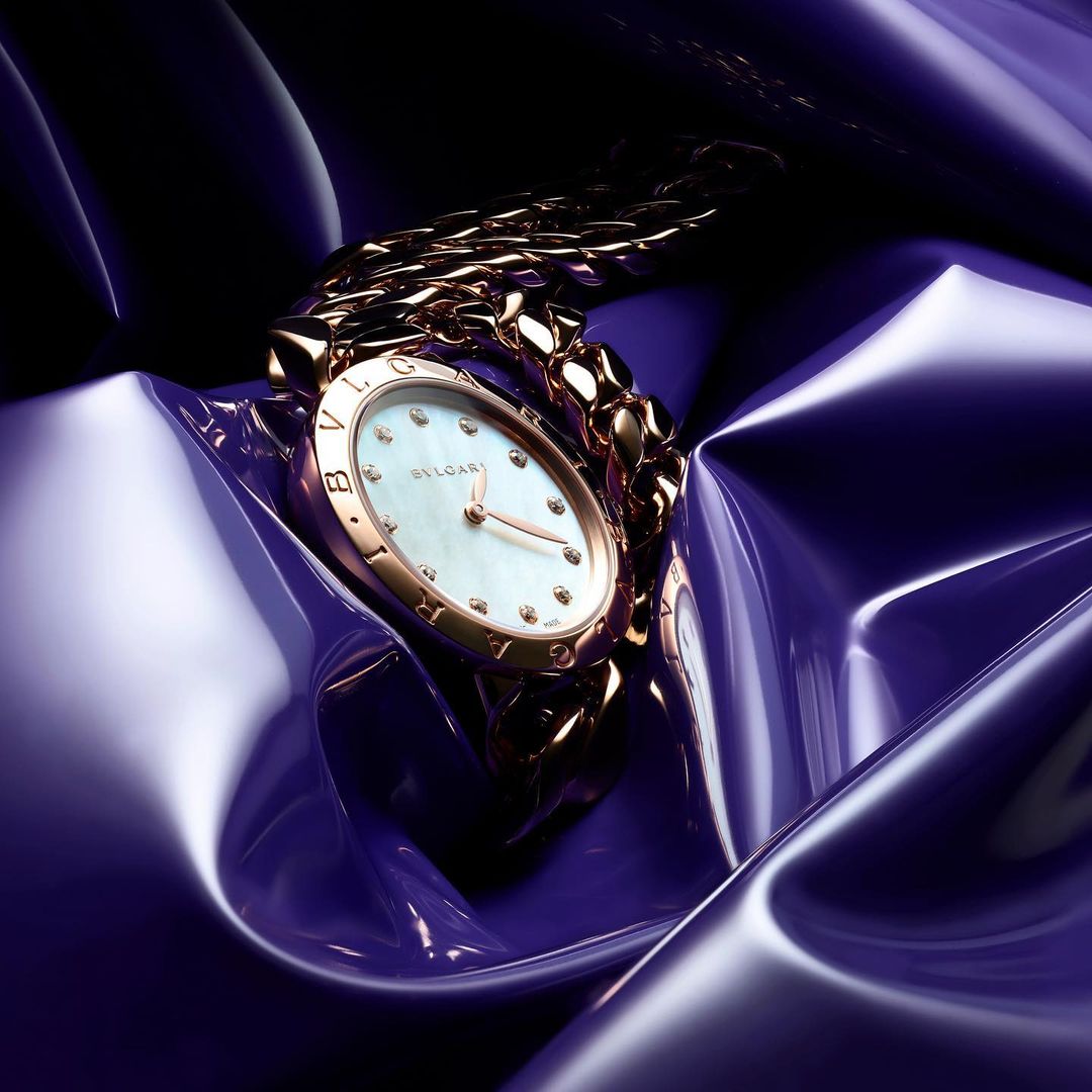 Close-up of a luxurious gold wristwatch with a white face and diamond accents resting on a shiny, textured purple fabric. Wristwatch is the focus.