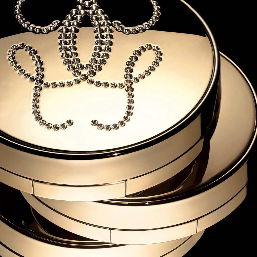 Close-up studio shot of stacked, round, shiny, gold-tone makeup compacts adorned with crystal detailing against a dark background.