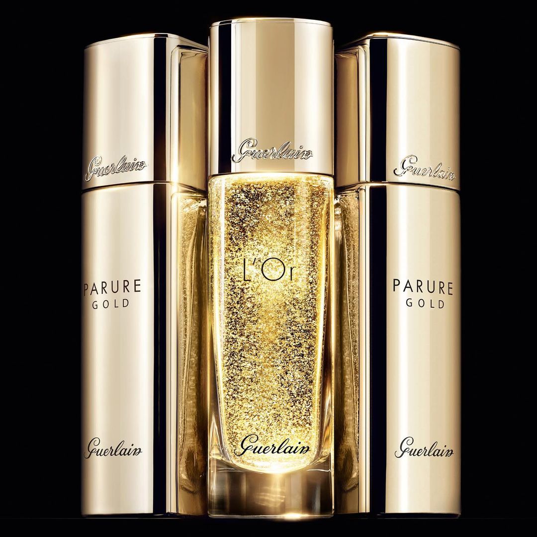 Guerlain Parure Gold skincare products in gold packaging displayed against a black background, close-up product shot. Gold liquid detail.