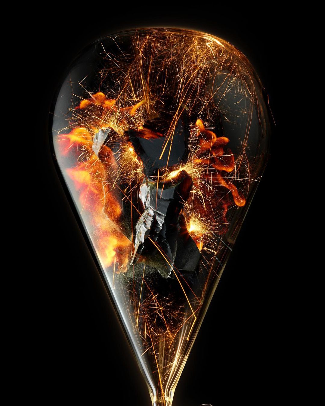 A glass vessel resembling a lightbulb contains fragments, possibly charcoal, surrounded by bright sparks against a black background.