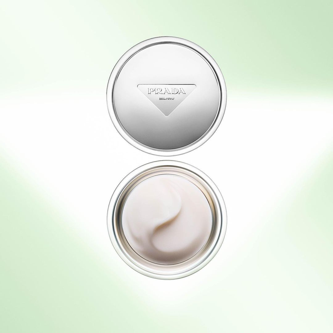 Overhead shot of open silver Prada skincare cream jar revealing white cream, displayed on a bright, soft green and white gradient background.