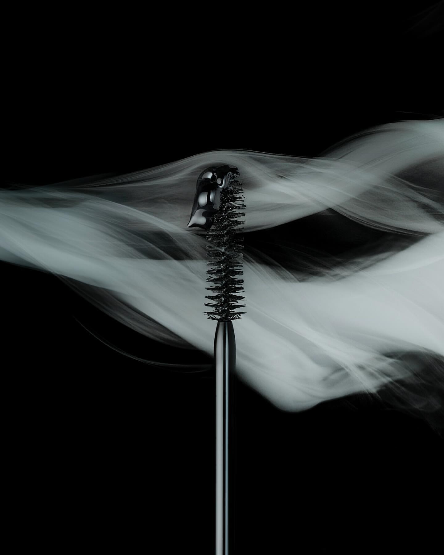 Close-up of a black mascara brush against a dark background with wisps of white smoke, showcasing beauty and cosmetic product.