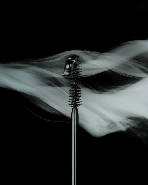 Close-up of a black mascara brush against a dark background with wisps of white smoke, showcasing beauty and cosmetic product.