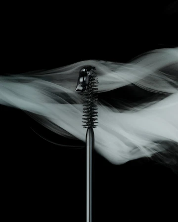 Close-up of a black mascara brush against a dark background with wisps of white smoke, showcasing beauty and cosmetic product.