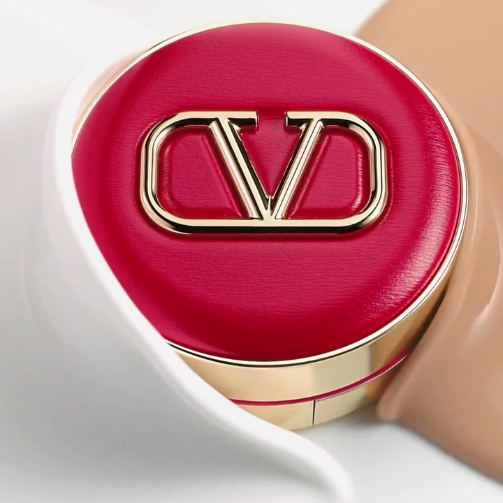 Close-up studio shot of a Valentino beauty compact with a red lid and gold logo, cream and beige product smears surrounding.