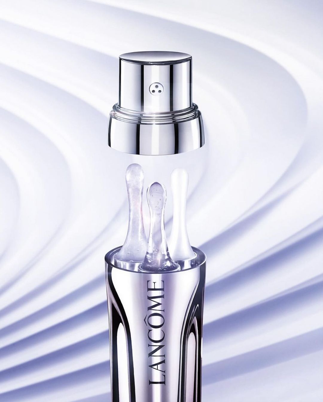Close-up product shot of a silver Lancôme cosmetics bottle with three applicator tips against a pale purple swirling background