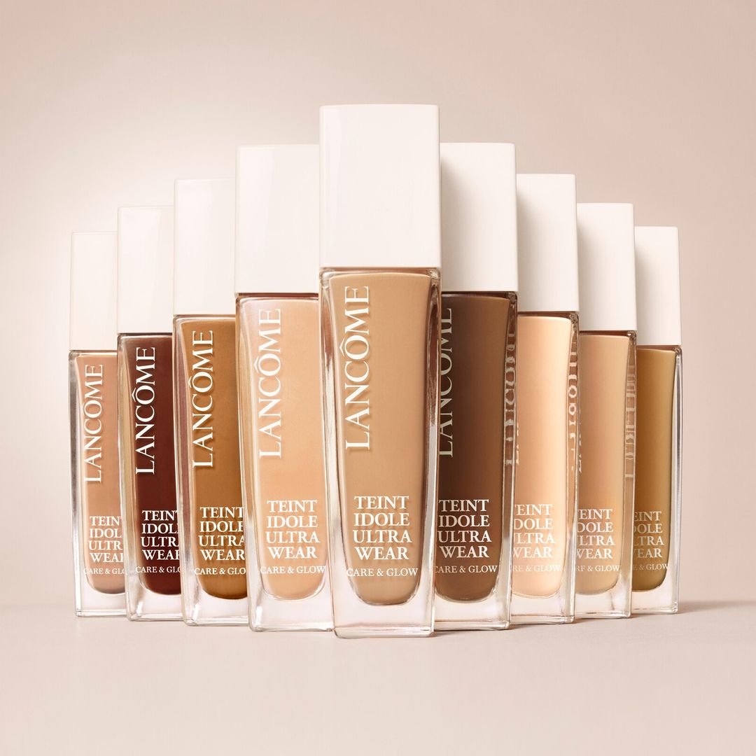 Close-up studio shot of an array of Lancome Teint Idole Ultra Wear foundation bottles with different skin tone shades against a beige background.