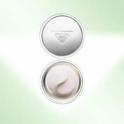 Prada skincare cream jar shown open with white cream and closed with silver lid against a light green gradient background.