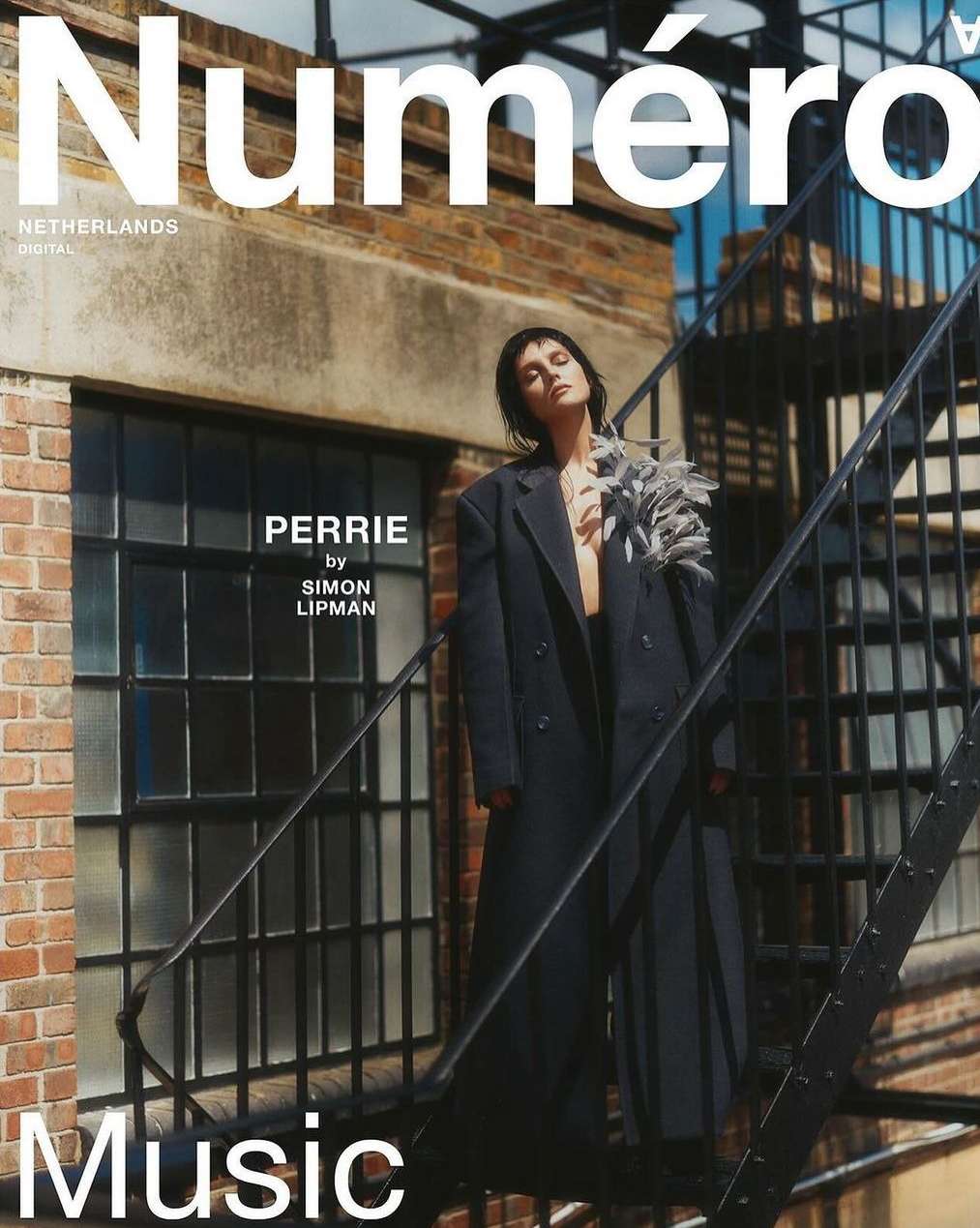 Magazine cover featuring a stylish woman in a black coat, standing against an urban backdrop with brick walls and stairs under bright sunlight.
