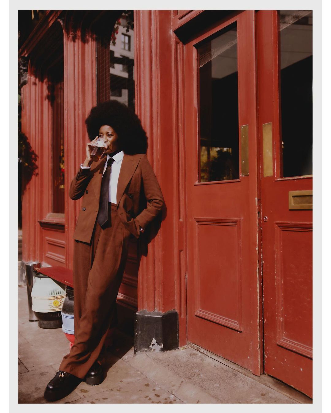 Stylish adult woman with Afro, wearing a brown suit and tie, drinks from a glass near a brown building exterior with a door.