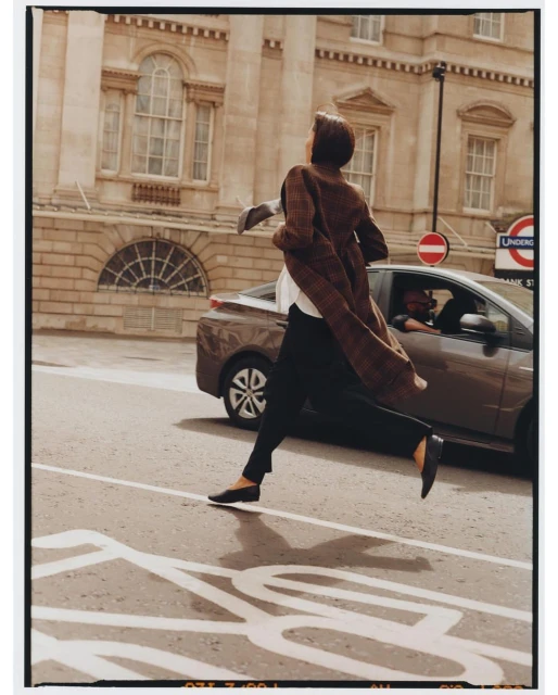 Woman in coat runs across urban street with a car and building in background, capturing a dynamic fashion editorial scene.