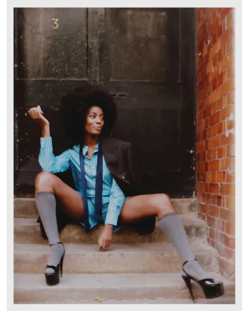 An African-American woman with an afro sits on outdoor steps, wearing a blue striped shirt, tie, blazer, socks and black platform heels.