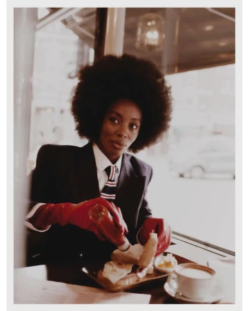 Black woman with afro wearing a suit jacket and red gloves squeezes ketchup onto a meal at a café table.