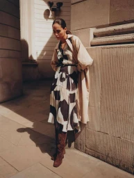 Model in patterned dress and beige trench coat leaning against a stone building by Simon Lipman