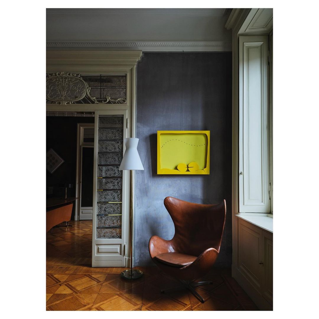 Interior with a tan leather chair, a floor lamp, and yellow art, in a room with dark grey walls and herringbone wood floors.