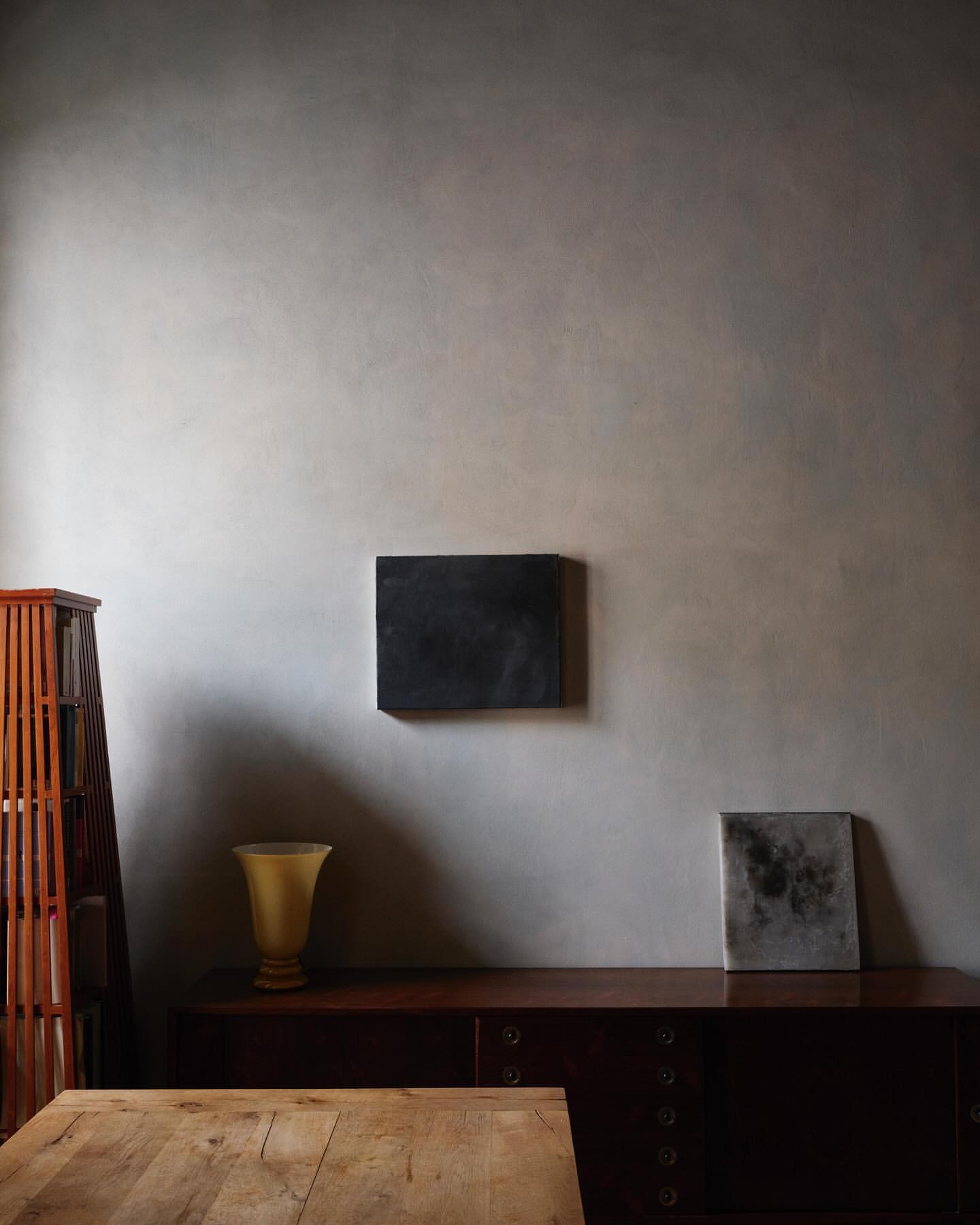 Interior view of a room featuring a wooden table, brown dresser, and wall art in shades of brown, grey and yellow with natural lighting.
