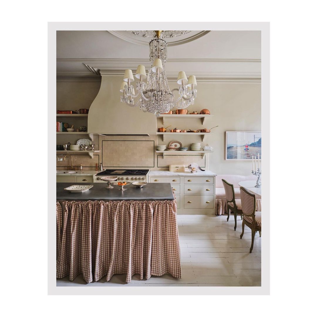 Traditional kitchen interior with island covered by checkered pattern fabric, crystal chandelier, wood floors, and beige walls.