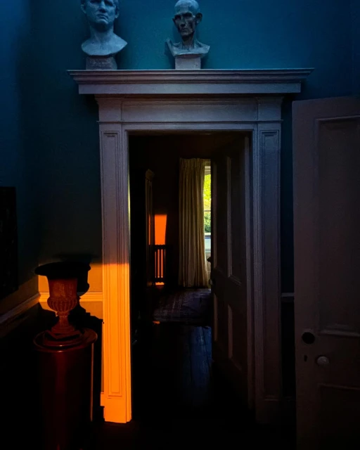 Interior view of an open doorway with natural light streaming in, illuminating busts on a mantelpiece and an urn in the foreground.