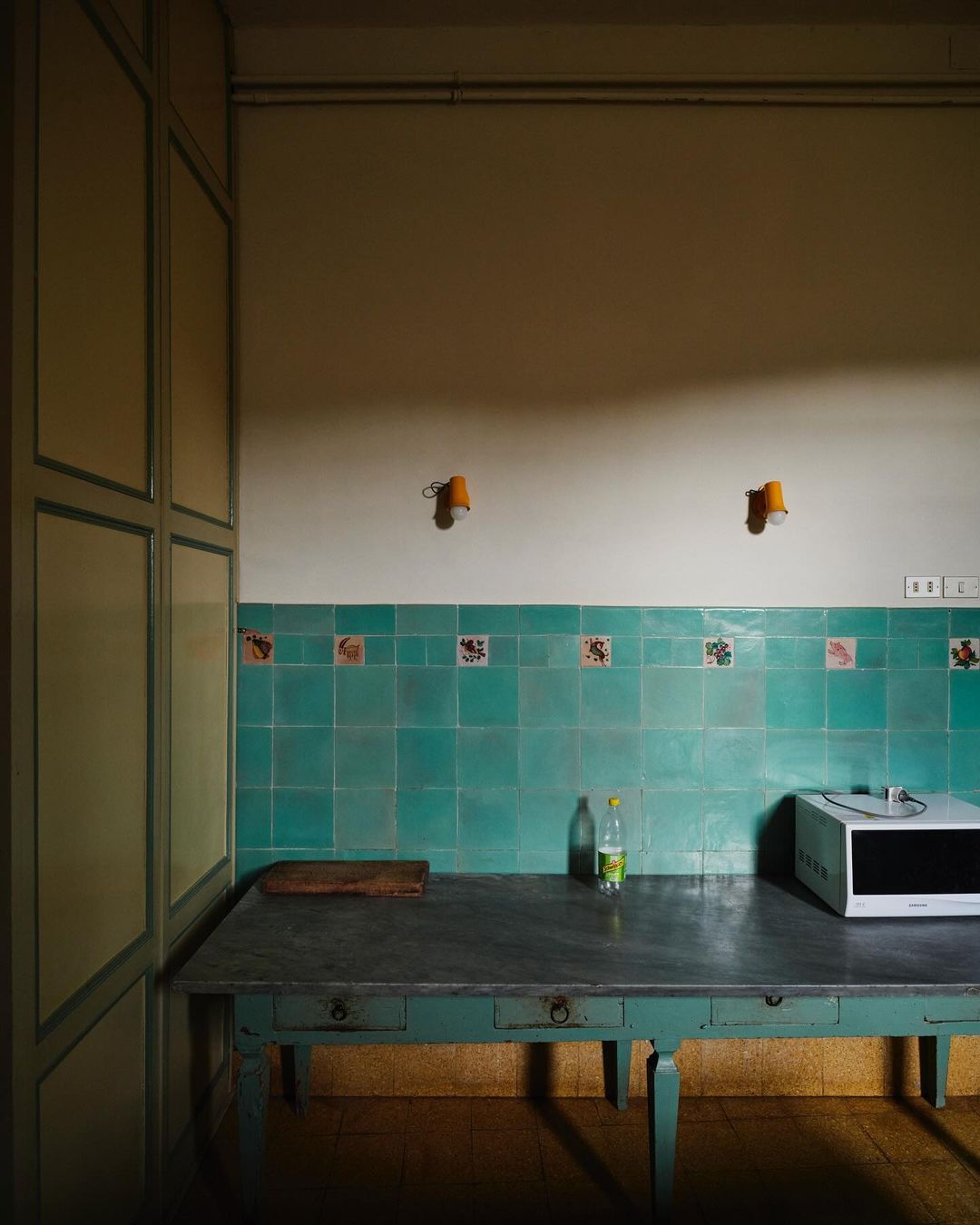 Interior shot of a vintage kitchen with teal tiles, pale yellow walls, and a weathered turquoise table displaying a bottle and microwave.