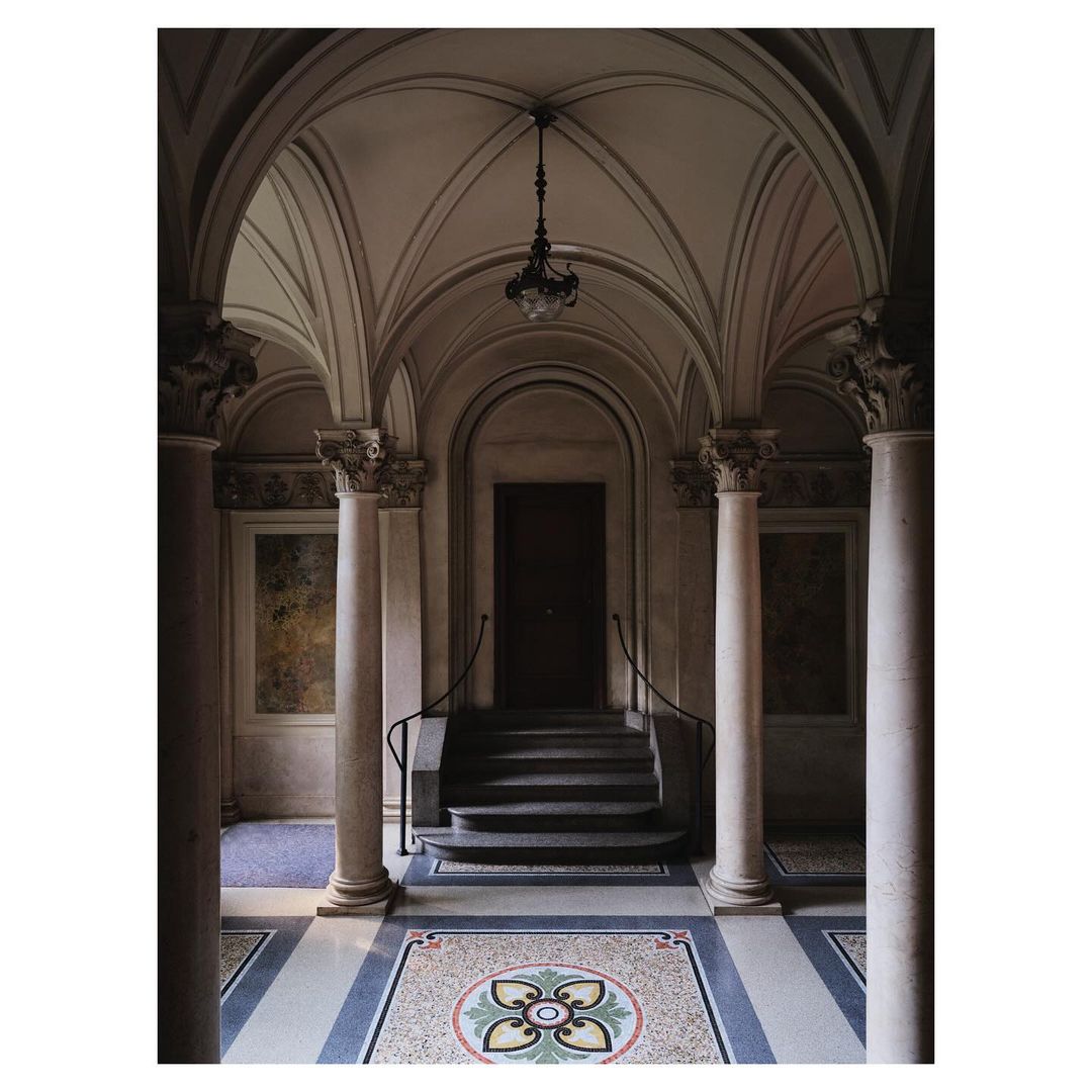 Elegant corridor with mosaic floor, arched ceiling, and classical columns leading to a dark wooden door and ornate hanging light.