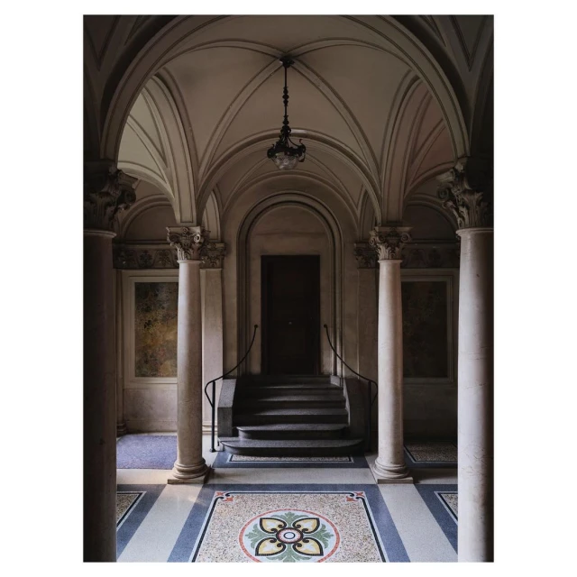 Elegant corridor with mosaic floor, arched ceiling, and classical columns leading to a dark wooden door and ornate hanging light.