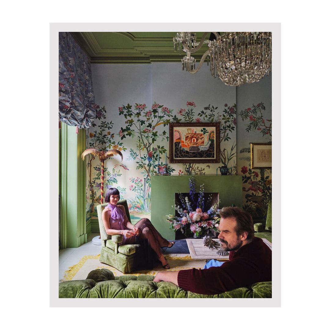 Couple seated in an elaborately decorated living room with floral wallpaper, vintage furniture, and a crystal chandelier.