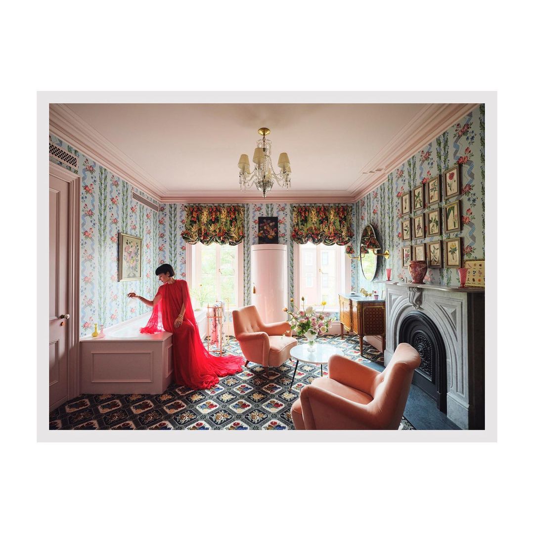 Woman in red gown poses in a luxurious room featuring floral wallpaper, elegant seating, and a vintage fireplace in an interior design shot.