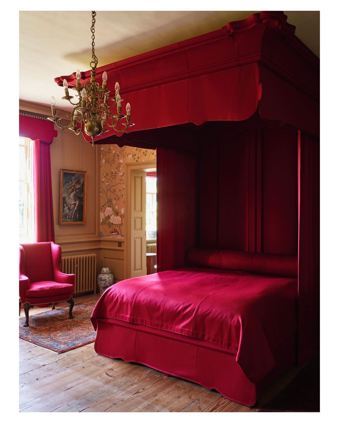 Luxurious four-poster bed dominates a warmly lit interior, complemented by an antique chandelier, classic armchair, and floral wallpaper.