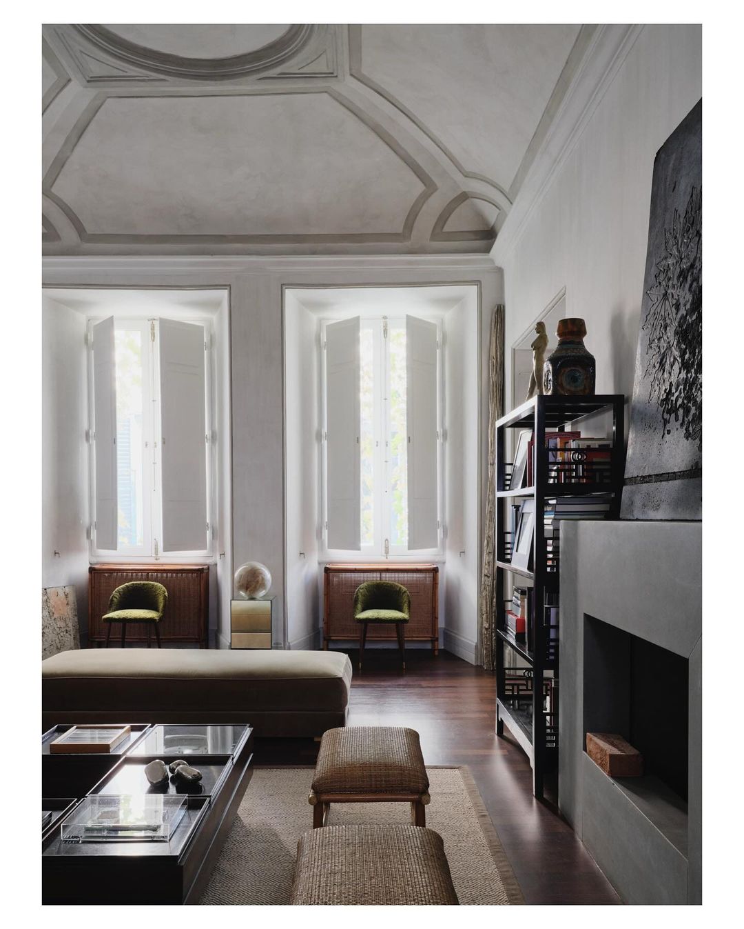 Living room interior with high ceilings, large windows with white shutters, a coffee table, ottomans, bookshelves, and gray walls.