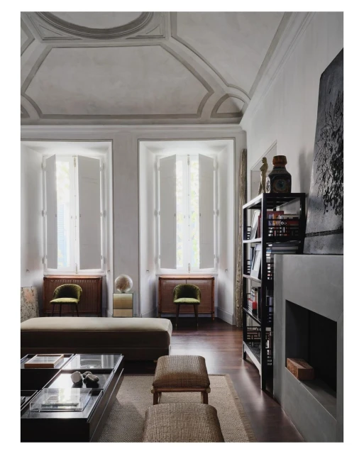 Living room interior with high ceilings, large windows with white shutters, a coffee table, ottomans, bookshelves, and gray walls.